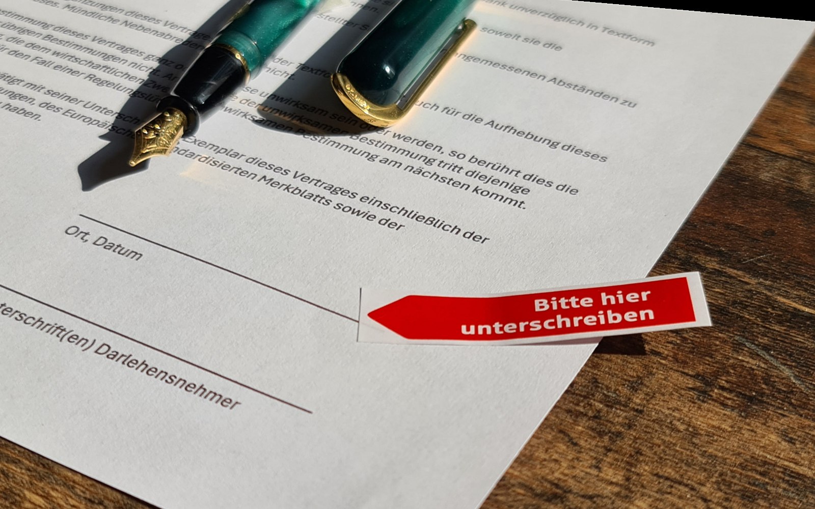 Ein Vertrag auf einem Holztisch, daneben ein Füllfederhalter mit goldener Feder; ein roter Marker mit der Aufschrift „Bitte hier unterschreiben“ weist auf die Unterschriftszeile